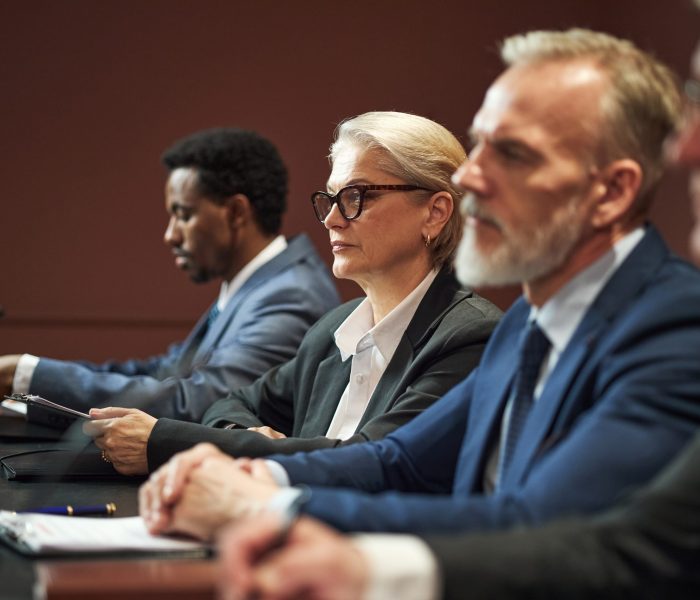 Middle aged Caucasian woman and middle aged Caucasian man sitting at conference table with Black man and another man, participating in political meeting, microphones visible